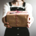 A woman holds a wrapped gift box, her face not visible, conveying a sense of anticipation and surprise.